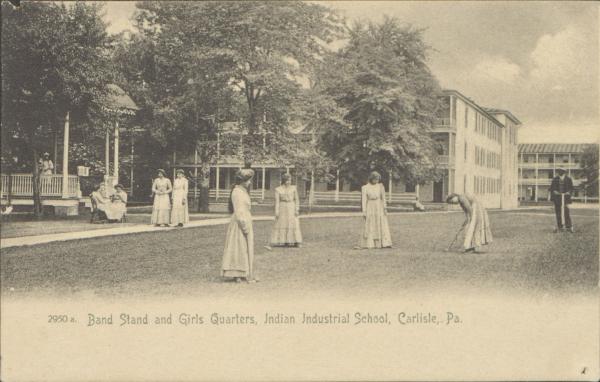 black and white image; in the foreground a group of young women are posed playing croquet, in the middle ground other young women sit on a bench by the band stand or walk, there is also a man with a lawnmower to the right, in the background is the bandstand, girls' quarters and just visible are the gymnasium and large boy's quarters