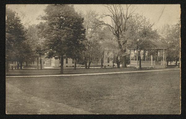 black and white image; view of the band stand and dining hall, trees obscure most of the dining hall from view, a path crosses in front of the band stand in the middle ground