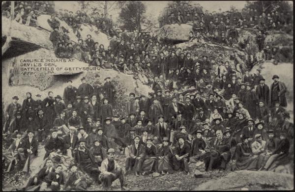 black and white image; a large group of students in dark clothing sit and stand around rocky outcropping