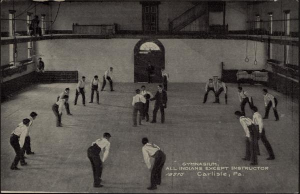 black and white image; a group of male students circled around two students and an instructor, they are poised as if ready to run, the students at the center seem to wait for instructor to give them a ball; photograph taken from the vantage point of the track