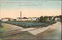 color image; view of campus looking southeast from the new entrance, road over water is in the foreground to the right, in the background to the left is the [smokestack] and surrounding buildings, in the background to the right is the housing for staff