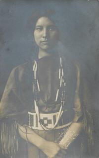 A real photo postcard, portrait of a young woman with two braids, she probably wears regalia. She is looking directly at the camera, hands clasped in front of her
