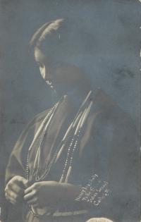 a real photo postcard, portrait of a young woman taken at a 3/4 view of her face, she looks down at something in her hands and seems to wear regalia