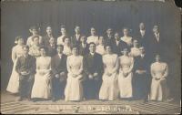 black and white image of 26 young people posed in three rows, the young women wear white dresses and white bows, the young men where black suits with various colored ties