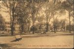 sepia-toned image; view of the band stand with the teacher's quarters in the background and a bench in the foregound, students sit on benches closer to the band stand