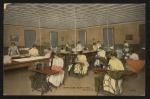 color image; in the foreground five young women sit at sewing machines, in the background a larger group of students stand at table cutting material, the background walls (intersected with five windows covered by blinds) are mustard yellow, most young women are dress in white or yellow dresses, cloth in hand of one of students at the sewing machine looks peach-colored