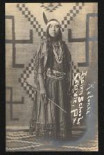 sepia toned portrait of a young woman in regalia, she is standing in front of a geometric-patterned wall hanging and holds a bow and arrow