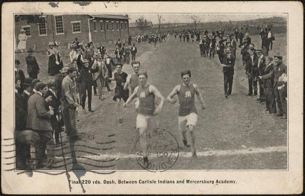 black and white image; photograph of the finish line of a track event, four young men are running towards the camera, the two in the front have a large M visible on their tops, a crowd of spectators stands on either side of the path to the finish line