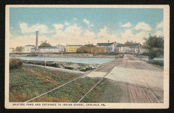 color image; view of campus looking southeast from the new entrance, road over water is in the foreground to the right, in the background is the smokestack, art studio, staff housing, a trolley is also in background on the tracks