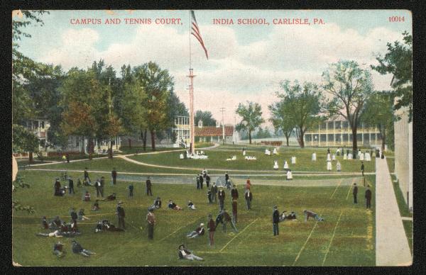 color image; male students lounging in foreground with women in the middle ground, guard house and academic building in the background in the middle and to the right respectively