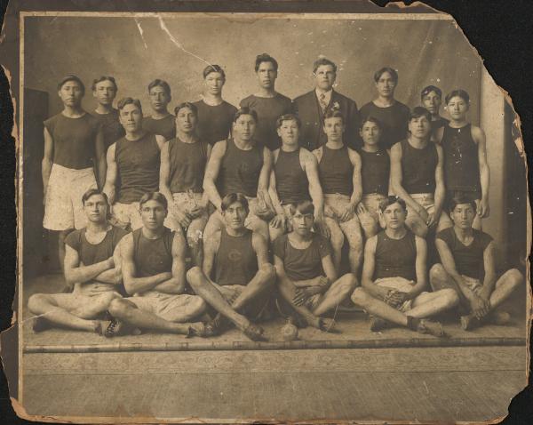 sepia toned photograph of 22 men sitting and standing in rows, all except Glenn Warner wear a dark shirt and light shorts with track shoes, two hold rabbits