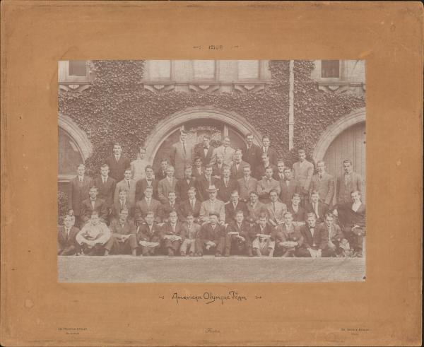 sepia toned photograph of 51 men sitting and standing in rows in front of ivy covered stonework