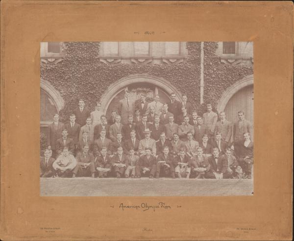 sepia toned photograph of 51 men sitting and standing in rows in front of ivy covered stonework