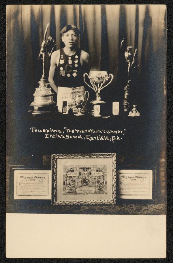 real photo postcard, portrait of Lewis Tewanima standing in front of a curtain and behind a table full of trophies. He wears an athletic uniform with a "c" on the front and six medals pinned on the front. Certificates for the 1908 Olympic Games sit in front of the table.
