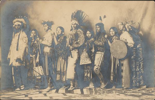 real photo postcard; group portrait of nine people standing in a row wearing regalia, two hold shields 