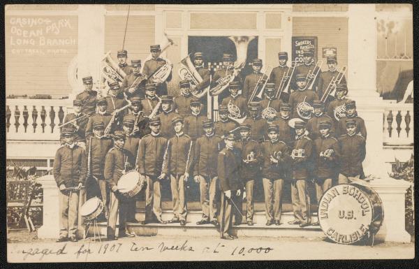 black and white image; group portrait of approximately 39 members of the Carlisle Indian School band and the band leader. The students stand in rows on the steps of a large building, in uniform, with instruments in hand