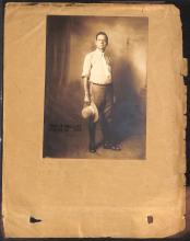 photograph of man standing in front of background in white short and brown pants, holding a hat