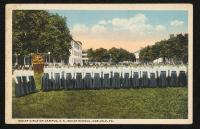 color image; in the foreground a large group of young women are stood in two rows, a banner which reads "Carlisle Indian School Competitive Drill" is at the left end of the row, in the background is the northeast end of campus (girls quarters, large boys quarters, small boys' quarters, trees); 