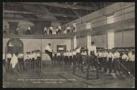 black and white image; male students in gymnasium, two using gymnastics equipment with a group of other students watching, also a set of students running in the track in the background