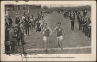 black and white image; photograph of the finish line of a track event, four young men are running towards the camera, the two in the front have a large M visible on their tops, a crowd of spectators stands on either side of the path to the finish line
