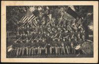 black and white image; photograph of the Carlisle Indian School band, fifty people sat on four levels of risers hold instruments, a bass drum with the words "Carlisle Indian School U. S." painted on it is visible in the right corner; 