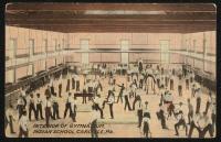 color image; view of the gynasium from the vantage point of the track, a large number of young men occupy the lower floor using equipment or watching those who are using the equipment; the rafters, wall and floor have been painted, the young men scattered throughout the room have not