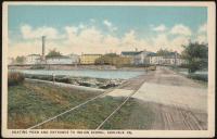 color image; view of campus looking southeast from the new entrance, road over water is in the foreground to the right, in the background is the smokestack, art studio, staff housing, a trolley is also in background on the tracks
