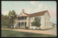 color image; view of dining hall (two story building with a small porch and two doors) from a diagonal angle, there is a road in front of the building and two trees (no people on porch)