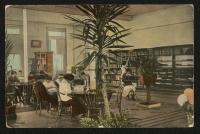 color image; view of interior of the library, a large plant sits in the foreground, in the middleground (to the left) are three tables of students looking down at study materials, in the background to the right are shelves of books and to the left is a window and door
