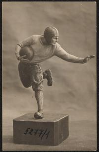 black and white image; photograph of a figure of a man running with a ball, he is wearing a head-covering, the ball is tucked into his right arm and his left arm is held out as if to fend something off