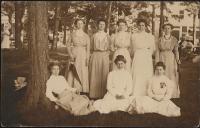 real photo postcard; portrait of eight young women, five standing and three sitting on the group, they are all wearing long dresses (most white) and have their hair up, one holds a penant that reads "Put-in-Bay," trees occupy the background