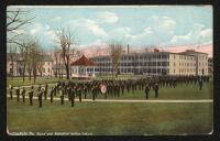 color image; a large group of male students stand on the green in front of the band stand, the front set carry instruments, in the background in the dining hall and girls' quarters; reproduced (and cropped) from photograph circa 1907
