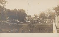 real photo postcard, view of section of school campus, male students lounge in the foreground on the tennis courts, female students are scattered in teh middle ground and around the flag pole, in the background is the guard house (to the left) and academic building (to the right)