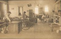 real photo postcard, view of six young men in a workshop, two stand near a set of wheels, two are in the background, and two stand to the right painting something