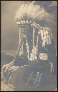 sepia-toned portrait of a young man sitting on the ground, he wears regalia and is lighting a pipe