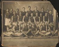sepia toned photograph of 22 men sitting and standing in rows, all except Glenn Warner wear a dark shirt and light shorts with track shoes, two hold rabbits