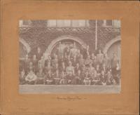 sepia toned photograph of 51 men sitting and standing in rows in front of ivy covered stonework