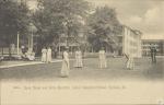 black and white image; in the foreground a group of young women are posed playing croquet, in the middle ground other young women sit on a bench by the band stand or walk, there is also a man with a lawnmower to the right, in the background is the bandstand, girls' quarters and just visible are the gymnasium and large boy's quarters