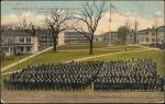 color image; view of the school grounds from perspective of the academic building towards the quarters, the trees have no leaves, in the foreground is a set of students arranged in tight rows (they are in black and white)