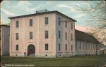 color image; view of the gynasium with one man standing by the entrance, appears faintly pink; the girls' quarters are just visible in the left corner of the image, the gymnasium is a three story building with three windows on each floor