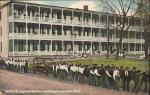 color image; in foreground a large group of young men pull equipment, in the background is the large girls quarters with a few young women and girls on the porch and nearby