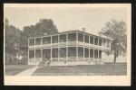 black and white image; a view of the adminsitration building with the Superintendent's Quarters just barely visible along the left side, building is two stories tall with a wrap-around porch on each level with white railings