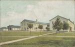 color image; view of the trade shops building (white painted two story building with blue roof, shaped like the letter u), small trees set along a path are in the middleground