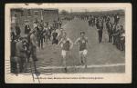 black and white image; photograph of the finish line of a track event, four young men are running towards the camera, the two in the front have a large M visible on their tops, a crowd of spectators stands on either side of the path to the finish line