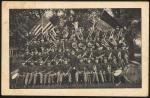 black and white image; photograph of the Carlisle Indian School band, fifty people sat on four levels of risers hold instruments, a bass drum with the words "Carlisle Indian School U. S." painted on it is visible in the right corner; 