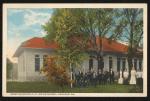 color image; view of the printing office (one story building with orange shingled roof), in front of the building is a set of trees and a small group of people (likely students and staff who worked in the printing office)