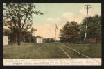 color image; view of campus looking northeast from the academic building, in the foreground are paint lines for tennis courts, in the middle ground is a fire hydrant, a tree to the left, and a telephone pole to the right, in the background is the gynasium, girls' quarters and large boys' quarters
