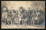 Nez Perce Students (pose 1) black and white image of nine young people in a photo studio, all sitting in one row with a cloth backdrop, they wear regalia, the person in the middle has a hand drum