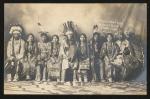 black and white image of nine young people in a photo studio, all sitting in one row with a cloth backdrop, they wear regalia, the person in the middle has a hand drum