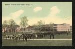 color image; in the foreground is the school band and behind them is a large group of male students, in the middle ground are trees and the bandstand, in the background are the dining hall, girls' quarters, and gymnasium